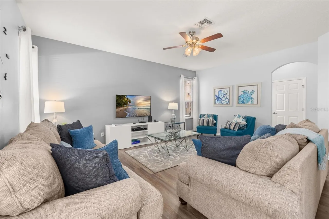 The great room: two deep sofas piled with blue cushions facing a wall-mounted TV, teal armchairs by the window and a ceiling fan turning overhead