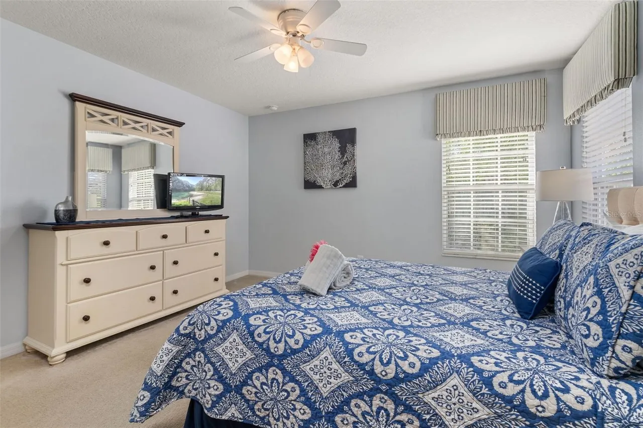 The primary suite: a king bed under a blue-and-white patterned quilt, a cream dresser with a mirror and TV, and plantation shutters on two windows